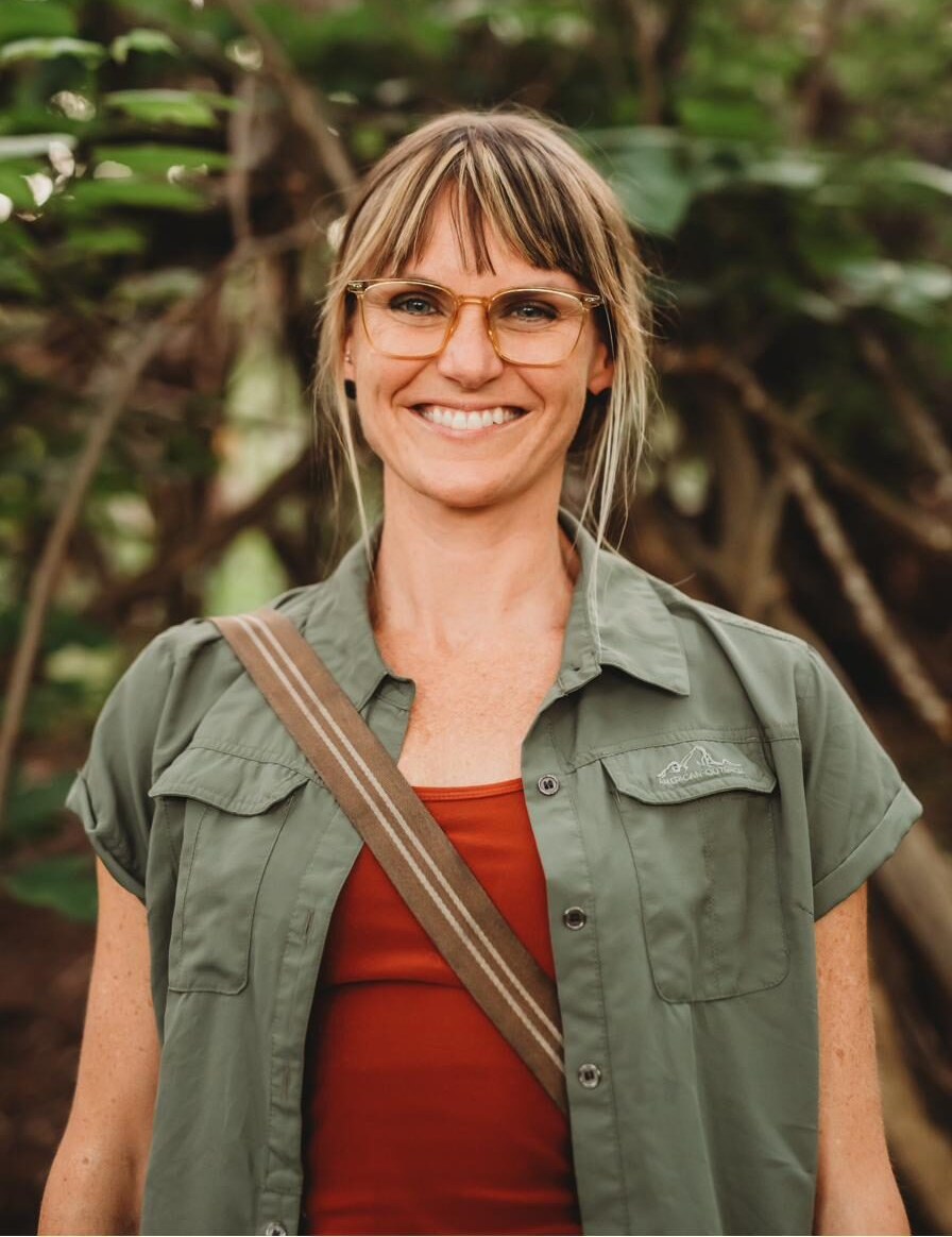 Summer Crider standing on a wooded trail in a green field shirt and rust tank top, a canvas bag strap across their shoulder, smiling at the camera.